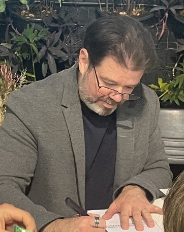 Alexandre Evangelista, homem de pele clara, barba grisalha e cabelos escuros, aparece sentado, concentrado enquanto autografa um livro. Ele usa óculos de leitura apoiados na ponta do nariz, blazer cinza e camiseta escura. Com a cabeça levemente inclinada para baixo, segura uma caneta preta na mão direita e escreve sobre papéis posicionados à sua frente. Ao fundo, há vasos e plantas ornamentais de folhas escuras, sugerindo um ambiente aconchegante, possivelmente externo ou em área de convivência. A cena transmite atenção e envolvimento durante o momento de autógrafos no 20º Encontro Internacional Phorte.
