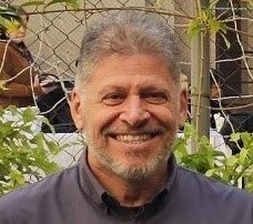 Professor Mario Charro, homem de pele clara, cabelos curtos e grisalhos e barba por fazer, sorri amplamente com os dentes à mostra. Ele veste camisa social cinza escura abotoada até o pescoço. Ao fundo, há uma placa preta de sinalização com ícones brancos indicando elevador e acessibilidade, sugerindo ambiente interno em área de circulação. O professor está parcialmente abraçado por alguém fora do enquadramento, em clima descontraído.