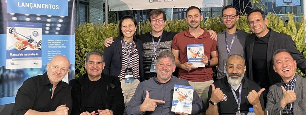 Grupo de dez pessoas posa sorrindo em frente a um banner azul com a palavra “LANÇAMENTOS” no topo, ao lado de um pôster com a capa do livro “Manual de Musculação: uma abordagem teórico-prática do treinamento de força”. Em ambiente externo com samambaias ao fundo, cinco pessoas estão sentadas e cinco estão em pé. Ao centro da fileira da frente, o professor Mario Charro sorri amplamente enquanto aponta para um exemplar do livro que segura. À sua direita, um homem barbudo faz sinal de positivo, e à sua esquerda, outro homem segura celular vermelho. Na ponta direita da fileira, está o professor Marco Uchida, de terno cinza claro, sorriso largo e olhar direcionado à câmera. Na fileira de trás, homens e mulheres, também com crachás do evento, celebram o lançamento do livro com expressões de alegria. A foto foi registrada durante o 20º Encontro Internacional Phorte.
