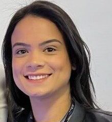 Luciana Capeloto, mulher jovem de pele clara e cabelos longos e lisos, pretos, aparece sorrindo suavemente para a câmera. Ela veste uma blusa escura com uma jaqueta preta por cima e usa um crachá com fita azul pendurado no pescoço, indicando participação no 20º Encontro Internacional Phorte. Ao fundo, a parede é branca e lisa, com iluminação clara. Alguns fios do cabelo estão levemente arrepiados no topo da cabeça, em uma pose espontânea e simpática.